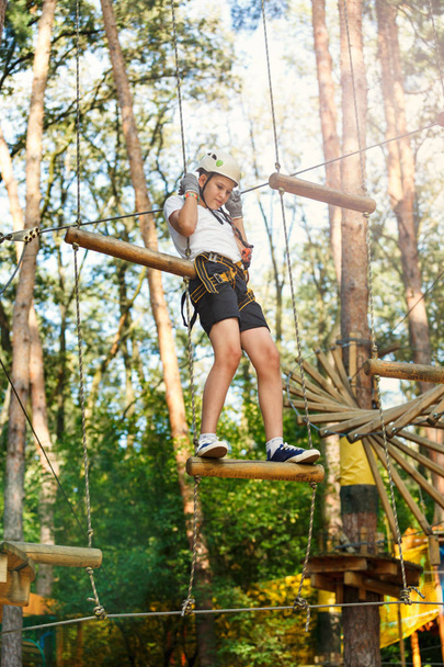 Cheerful cute young boy in white t shirt and white helmet in adventure rope park at sunny summer day. Active lifestyle, sport, holidays for children - Photo, Image