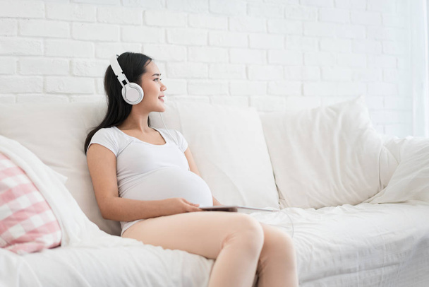 Portret van Aziatische zwangere vrouw zittend op de Bank thuis en luisteren muziek in hoofdtelefoons. - Foto, afbeelding