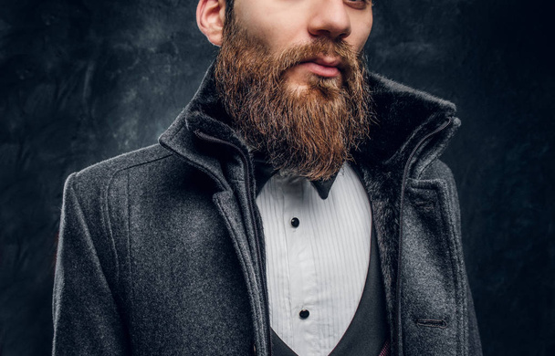 Imagen recortada de un hombre barbudo con estilo vestido con un elegante traje y abrigo de pie contra una pared de textura oscura
 - Foto, Imagen