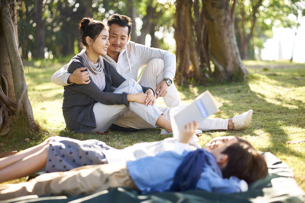 joven asiático pareja sentado en hierba en parque charlando con dos niños mintiendo lectura libro en primer plano
. - Foto, Imagen