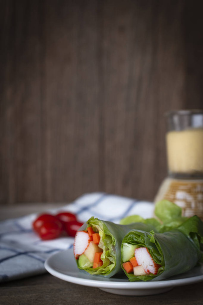 Salaattirullat valkoisella levyllä, joka on sijoitettu puupöydälle, jossa on valkoinen sininen raidallinen kangas, tomaatti ja salaattikastike takapuolelle
. - Valokuva, kuva