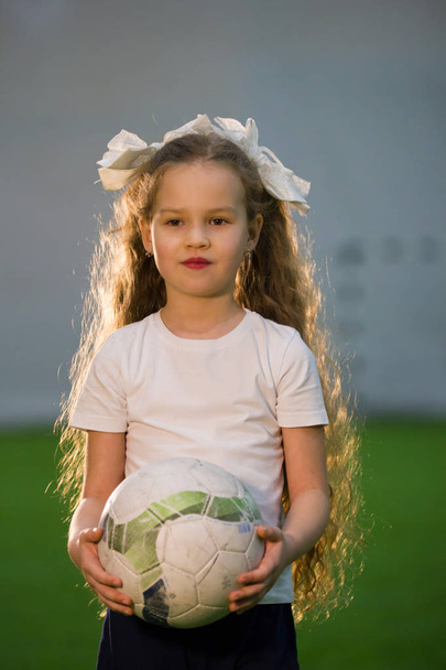 Ellerinin üzerinde futbol sahası içinde topu tutarak ve gülümseyen uzun sarı saçlı küçük bir kız - Fotoğraf, Görsel