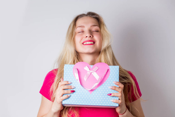 An attractive cute blonde woman received a gift for her birthday, Valentine's day. Joyful smiling girl holding a gift bag with a pink heart and closes her eyes on a white background - Photo, image