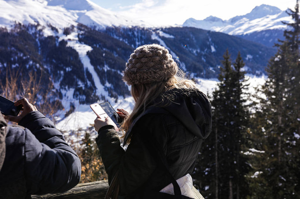 Tourist woman wearing warm clothes, standing on the observation deck and taking pictures of incredible nature beauties around. High powerful mountains and dense pine forest. A lot of white snow. - Photo, Image