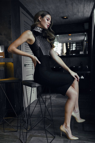 Young brunette model in cocktail black dress with open arms and high hell shoes sitting on the bar chair near the bar. Hand on the back, looking down. Volume curly hair lying on shoulder, makeup  - Valokuva, kuva