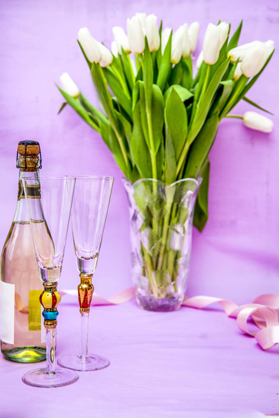 2 glasses, a bottle of sparkling rose wine and a vase with white tulips on a light purple background - Foto, imagen