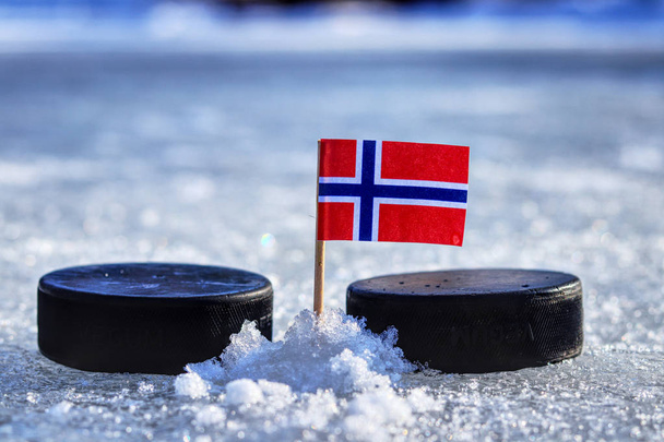 A Norwegian flag on toothpick between two hockey pucks on ice in outdoor. A Norway will playing on World cup in group B. 2019 IIHF World Championship in Slovakia. - Photo, Image