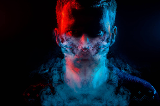 Retrato de un hombre que está enojado y tiene malas emociones con la máscara de un humo alrededor sobre un fondo negro aislado. La cara del tipo en la sombra con el color rojo y azul de la iluminación
. - Foto, Imagen