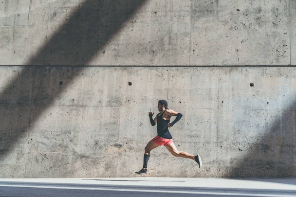 Gespierde mannelijke atleet sprinter snel uitgevoerd, uit te oefenen buiten, joggen buiten tegen grijze concret achtergrond met ruimte kopieergebied voor SMS-bericht of advertentie-inhoud. Zijaanzicht, volledige lengte - Foto, afbeelding