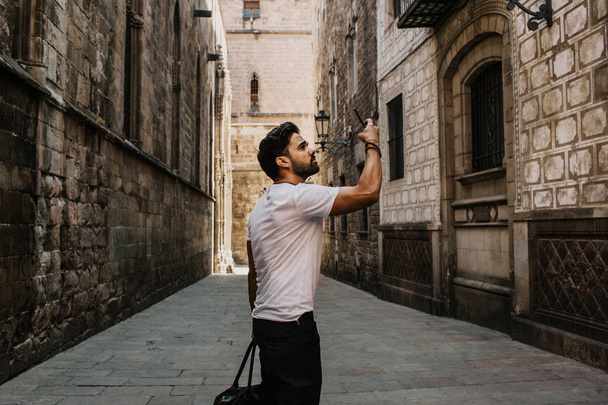 Content attractive hispanic man in white tshirt photographing himself on mobile phone while enjoying city travel.Bearded hipster male making selfie via smartphone on gotic streets. - Foto, Bild