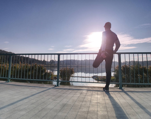 Lábak nyújtás előtt haladó fuss kívül belül napsütéses reggel ember. Bemelegítés az izmok. Szabadtéri tó hídon gyakorlása. - Fotó, kép