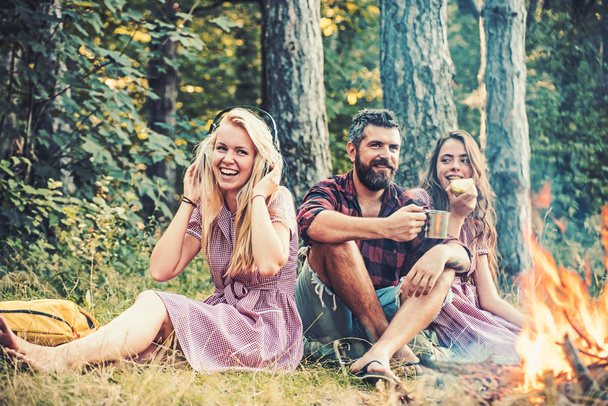 People camping at fire in forest in vintage style. Friends relax at bonfire flame with sparks. Women and bearded man at campfire. Eating food, reading book and entertainment. Summer vacation concept - Photo, Image