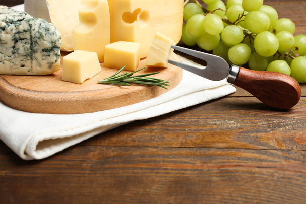 Cheese platter: yellow Maasdam cheese, white Camembert cheese and blue cheese Dor Blue with rosemary, grapes and fork on cutting board on wooden background. Copy space.  Concept serving cheese. - Photo, Image