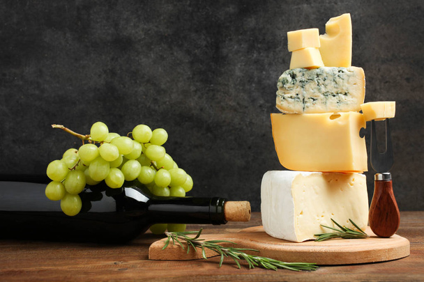 Πιατέλα τυριών: κίτρινα τυριά ελβετικο τυρι, λευκό τυρί Camembert και μπλε τυρί Dor μπλε με μπουκάλι κρασί και σταφύλια σε ξύλινο τραπέζι σε μαύρο φόντο. Έννοια μερίδα τυρί. - Φωτογραφία, εικόνα