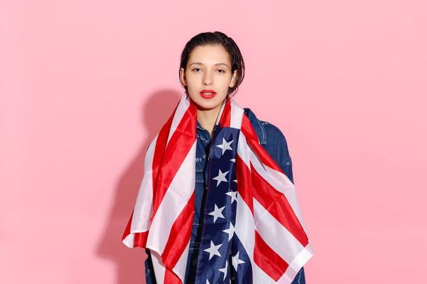 Portrait of proud female athlete wrapped in American Flag against pink background. Muscular young woman looking confidently at camera. female protesting for equality and women empowerment - Foto, Bild