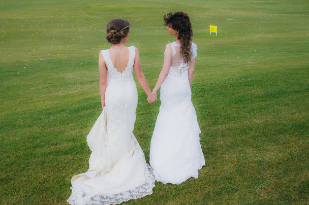 Duas belas noivas de mãos dadas no campo verde do taco de golfe
 - Foto, Imagem