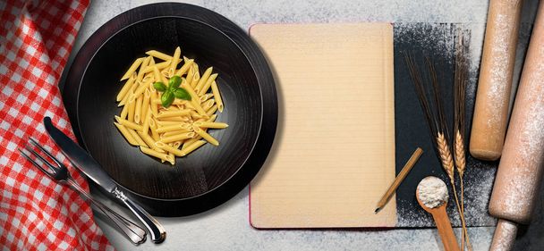 Italian pasta called Penne and an empty recipe book on a table with kitchen utensils, flour, rolling pin, ears of wheat, checkered tablecloth, fork and knife - Valokuva, kuva