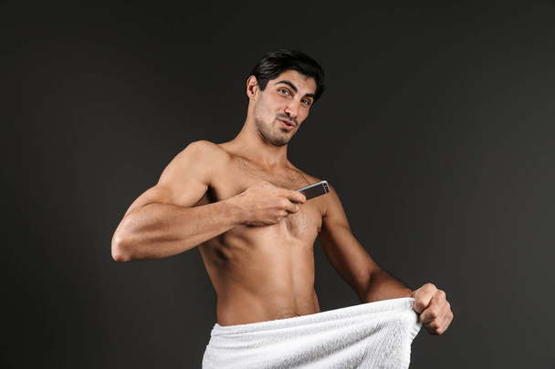 Image of a young naked man isolated over dark background covering his genitals with towel take an intimate photo by mobile phone. - Foto, Imagem