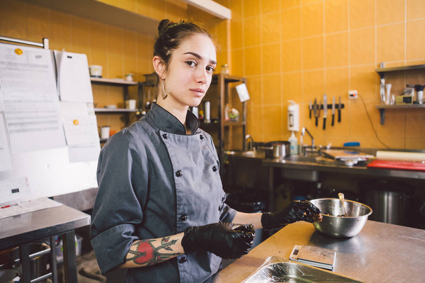 Onderworpen beroep en koken gebak. jonge Kaukasische vrouw met tatoeage van de chef van het gebakje in keuken van restaurant voorbereiding ronde chocolade snoepjes handgemaakte truffel in zwarte handschoenen en uniform. - Foto, afbeelding