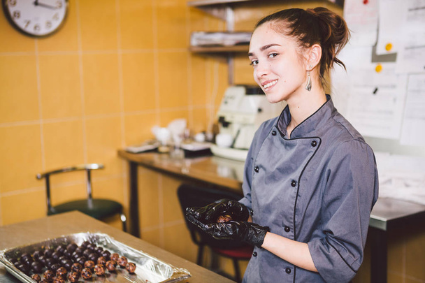 Subject profession and cooking pastry. young Caucasian woman with tattoo of pastry chef in kitchen of restaurant preparing round chocolate candies handmade truffle in black gloves and uniform. - Photo, Image