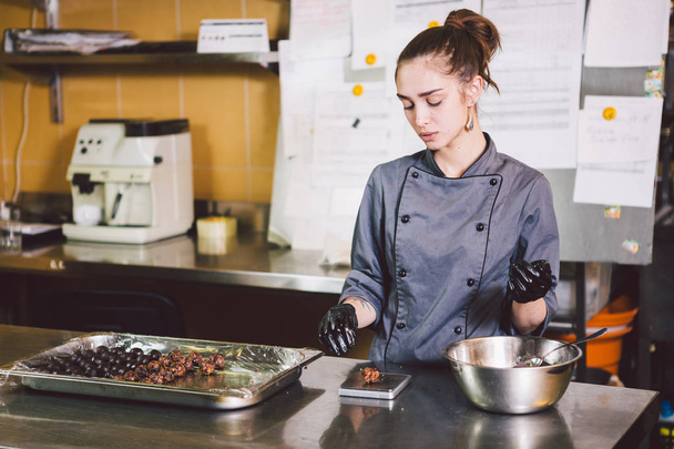 Subject profession and cooking pastry. young Caucasian woman with tattoo of pastry chef in kitchen of restaurant preparing round chocolate candies handmade truffle in black gloves and uniform. - Foto, afbeelding