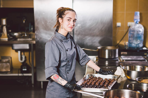 Subject profession and cooking pastry. young Caucasian woman with tattoo of pastry chef in kitchen of restaurant preparing round chocolate candies handmade truffle in black gloves and uniform. - Photo, Image