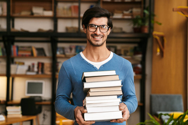 Przystojny młody mężczyzna noszenie okularów, stojąc w bibliotece, trzymając stos książek - Zdjęcie, obraz