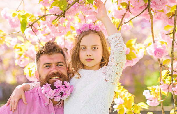 Girlish leisure concept. Girl with dad near sakura flowers on spring day. Child and man with tender pink flowers in beard, wreath on head. Father and daughter on happy faces hugs, sakura background - Foto, afbeelding