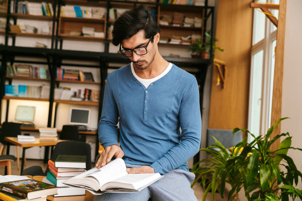 schöner junger Mann mit Brille steht in der Bibliothek und hält einen Stapel Bücher in der Hand - Foto, Bild