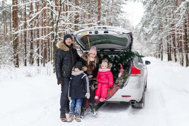 Μια ευτυχισμένη νεαρή οικογένεια ετοιμάζεται για τα Χριστούγεννα πρωί, στέκεται στο πίσω μέρος του αυτοκινήτου στο δάσος, χαμογελά στη φωτογραφική μηχανή. Οικογενειακό πορτρέτο - Φωτογραφία, εικόνα