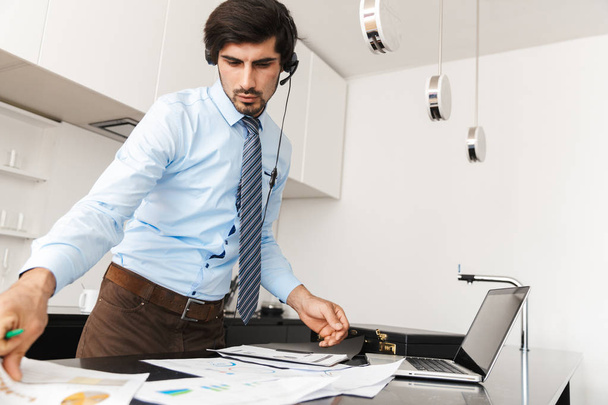 Image of a concentrated young business man at the kitchen wearing headphones work with documents. - Photo, image