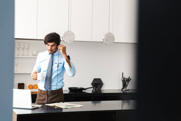 Confident young businessman working at home at the kitchen, using laptop computer, communicating through headphone set, drinking coffee - 写真・画像