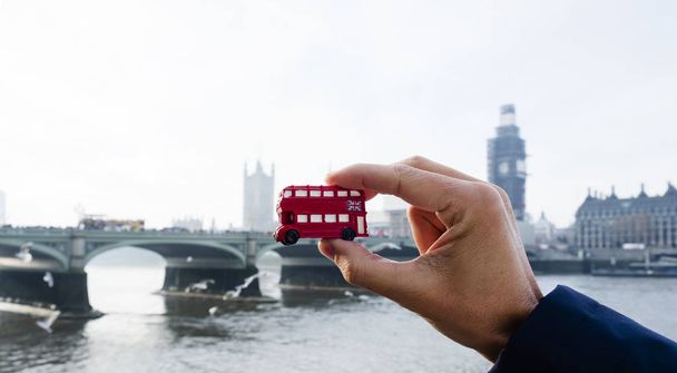 the hand of a caucasian man holding a miniature of a red double-decker bus, typical of London, United Kingdom, with the Westminster bridge and the the Palace of Westminster in the background - Photo, Image