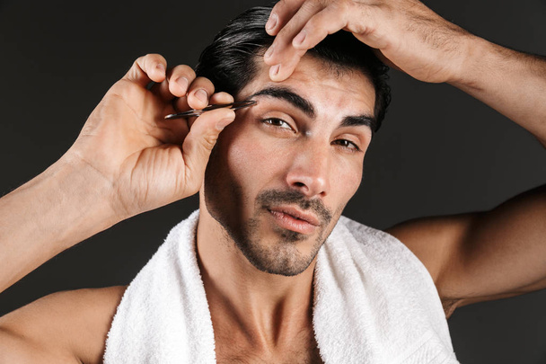 Image of a handsome young man posing isolated over dark wall background correct brows form with tweezer. - Φωτογραφία, εικόνα