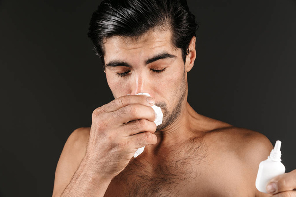 Imagen de un joven enfermo posando aislado sobre fondo de pared oscura sosteniendo botella de aerosol nasal con servilleta
. - Foto, imagen
