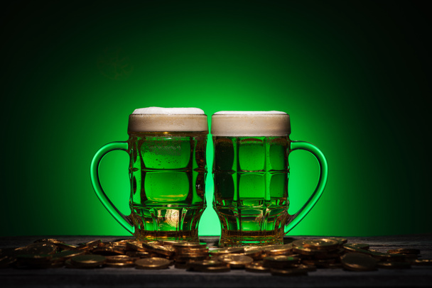 glasses of alcohol beer standing near golden coins on st patricks day on green background - Foto, Imagen