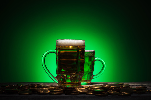 glasses of irish beer standing near golden coins on green background - Foto, Imagem