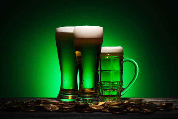 glasses of irish beer standing near golden coins on table on green background - Fotoğraf, Görsel