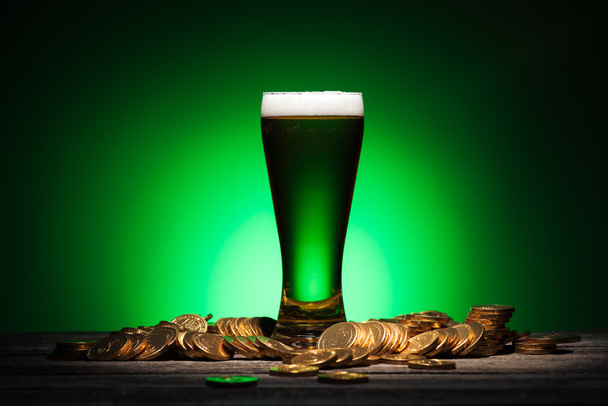vidro de cerveja irlandesa verde em pé sobre mesa de madeira perto de moedas no fundo verde
 - Foto, Imagem