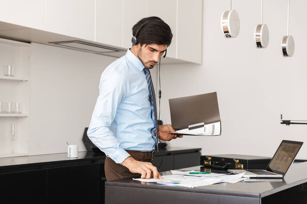 Image of a concentrated young business man at the kitchen wearing headphones work with documents. - Photo, Image