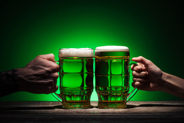 cropped view of friends holding glasses of beer on st patricks day on green background - Foto, Imagen