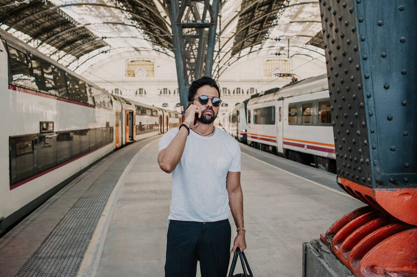 Confident positive bearded businessman wearing sunglasses standing with travel bag at the hall of railway station and using mobile phone. - Фото, зображення