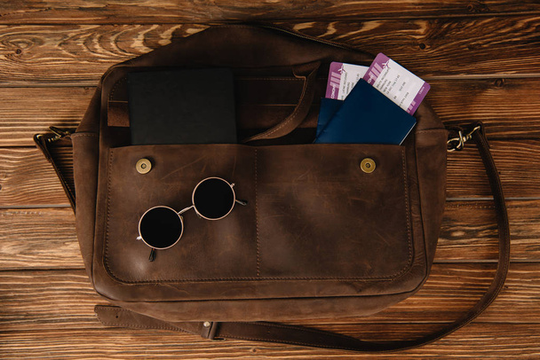 κάτοψη του καφέ δερμάτινη τσάντα με γυαλιά ηλίου και διαβατήρια με εισιτήρια - Φωτογραφία, εικόνα
