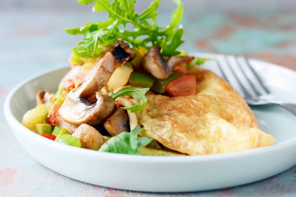 Homemade omelette with mushrooms, vegetables and arugula on a plate. Wonderful healthy breakfast near the window with a cup of coffee. - Photo, Image