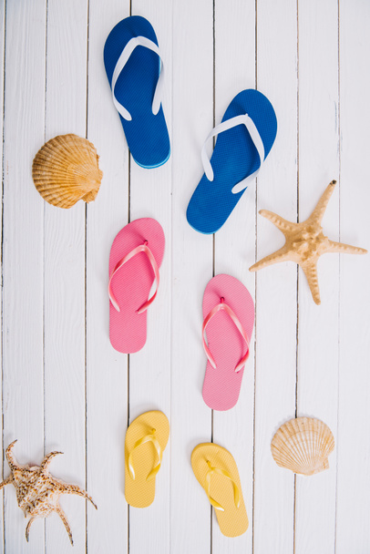 top view of yellow, blue and pink flip flops on white wooden background with seashells - Φωτογραφία, εικόνα