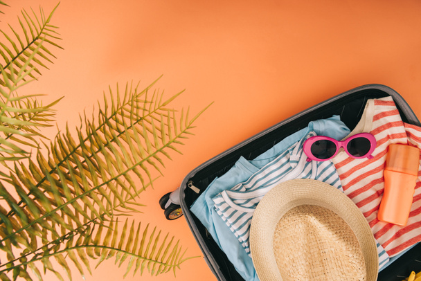 top view of suitcase with summer accessories and sunscreen on orange background with palm leaves - Foto, immagini