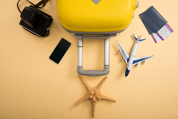 top view of yellow suitcase, plane model, starfish, film camera, smartphone and tickets on beige background - Foto, Imagem