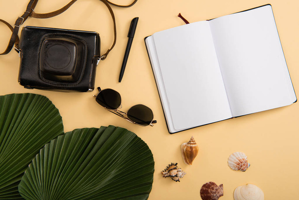 top view of empty notebook, film camera, seashells, palm leaves and sunglasses on beige background - Foto, Imagen
