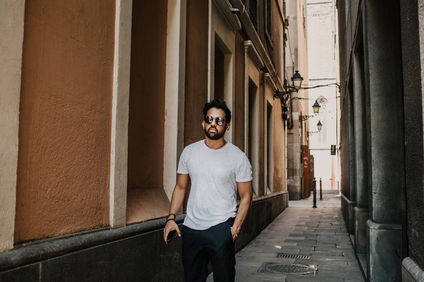 Content attractive hispanic man in white tshirt walking old town streets holding mobile phone hand.Bearded hipster male enjoying city travel on gotic district streets. - Valokuva, kuva