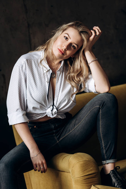 Portrait of a beautiful fashionable woman, blonde, jeans, on a yellow sofa - Foto, afbeelding